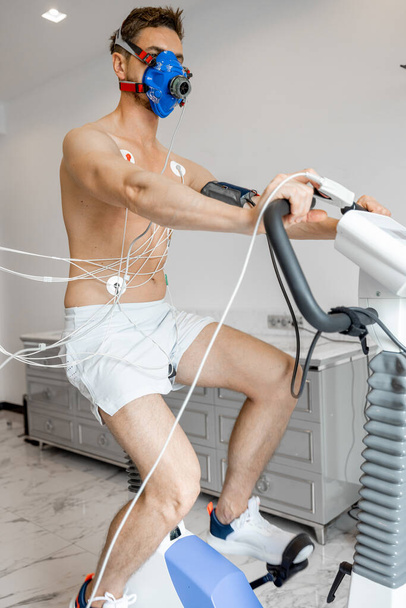 Man athlete with breath mask and electrodes training on bike simulator, examining his cardiovascular system at medical office - Foto, immagini