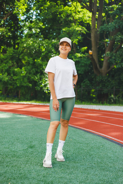 Μια νεαρή γυναίκα με λευκό μπλουζάκι και αθλητικά παπούτσια, ένα καπέλο στο κεφάλι της. Προθερμαίνεται πριν την προπόνηση στο στάδιο. Υγιής τρόπος ζωής - Φωτογραφία, εικόνα