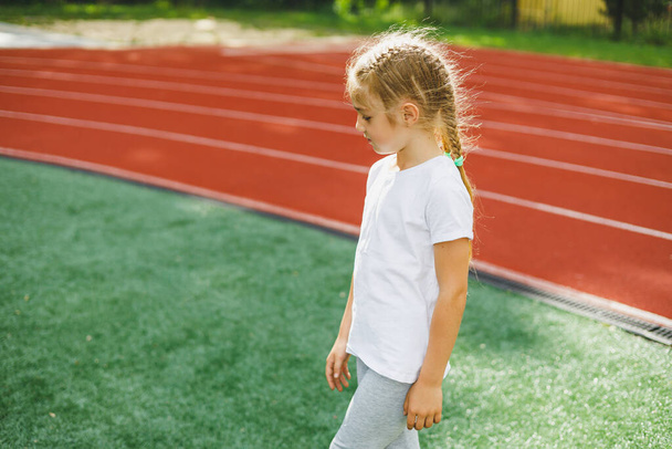 Mała dziewczynka w wieku 7-8 lat w białym t-shircie biegnie na zewnątrz na stadionie. Dziewczyna uprawia sport w słoneczny dzień. - Zdjęcie, obraz