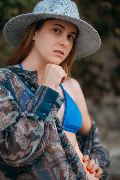 長いブロンドの髪とスポーティーな姿をした魅力的な若い女の子の肖像画,セクシーな青い水着で,小さな帽子と青い夏のシャツ,自然の中で立っています. - 写真・画像