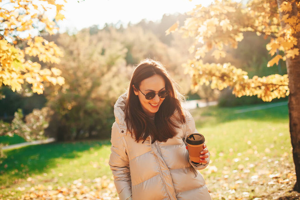 Smiling happy young woman with disposable coffee cup to go, enjoying walking in the sunny autumn park - Foto, Imagem