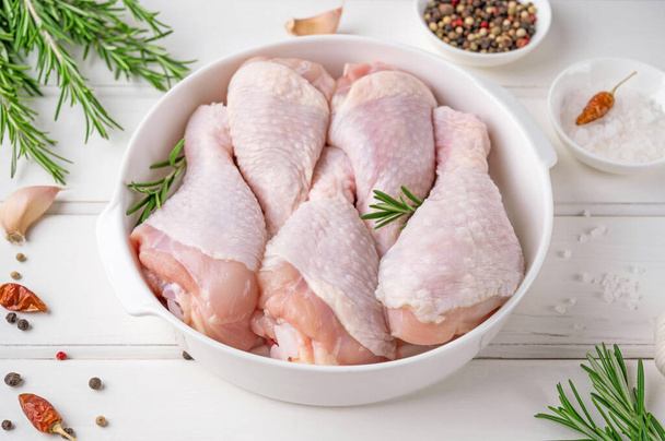 Pernas de frango crus não cozidas em um prato branco em um fundo de madeira branco. Carne com ingredientes para cozinhar. Vista superior - Foto, Imagem