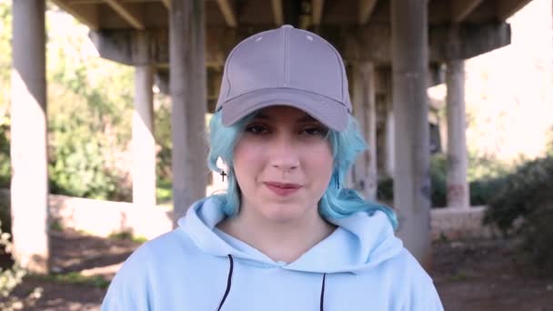 Una ragazzina sorridente con un cappello da baseball. Ragazza adolescente dai capelli blu che sta all'aperto contro i pilastri del ponte. Mockup, colpo alla testa. Concetto di hipster e adolescenza - Filmati, video