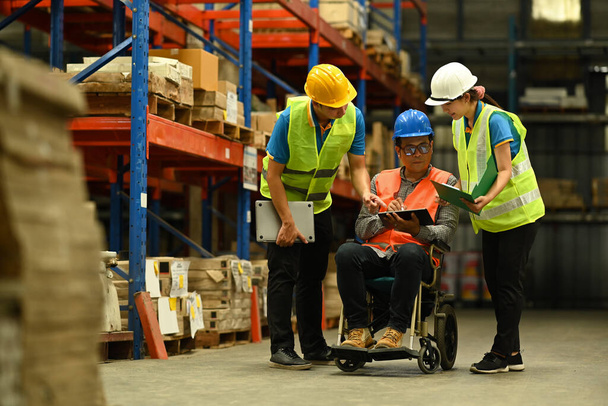 Ασιάτης διευθυντής σε αναπηρική καρέκλα και νέοι εργαζόμενοι που χρησιμοποιούν ψηφιακή ταμπλέτα, επιθεώρηση προϊόν αποθέματος σε μια μεγάλη αποθήκη. - Φωτογραφία, εικόνα