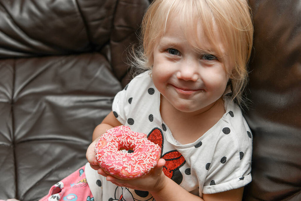 Ein kleines fröhliches Mädchen hält einen roten Donut mit Zuckerguss in ihren Händen. - Foto, Bild