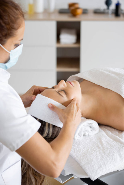 Обрізати жіночого масажиста в стерильній масці масажуючи обличчя клієнта із закритими очима на столі в салоні
 - Фото, зображення