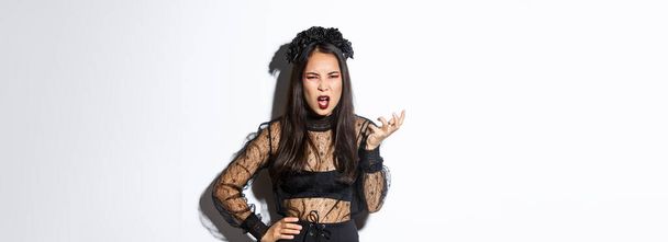 Frustrated and angry young asian woman in witch dress arguing with someone, looking pissed-off and bothered, shaking hand and looking scary at camera, standing over white background. - Foto, imagen
