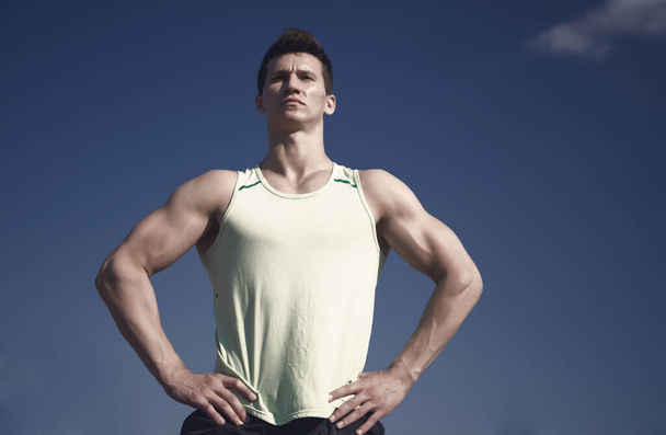 Athlete in green tshirt on sunny day. Man bodybuilder posing on blue sky. Sport and healthy lifestyle concept. Fitness model with muscular hands, biceps, triceps, strong muscles. Workout and training. - Valokuva, kuva