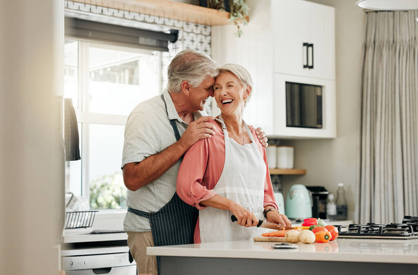 Idősebb pár a konyhában, egészséges ételeket főznek együtt, és boldogok a nyugdíjas életvitelben. Idős nő aprítás zöldségek kötény, öreg fehér férfi ölelés feleség otthon és szerelem táplálkozás vacsora. - Fotó, kép