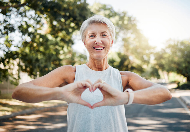 Fitness, happy and heart hands of old woman in nature after running for health, wellness and workout. Smile, motivation and peace with senior lady and sign for love, faith and training in nature. - Photo, Image