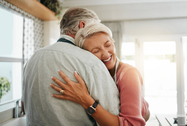 Αγκαλιά, αγάπη και ασφάλεια με ένα ζευγάρι ηλικιωμένων να αγκαλιάζονται ή να αγκαλιάζονται στην κουζίνα του σπιτιού τους μαζί. Ευτυχισμένος, χαμογελαστός και στοργικός με έναν ηλικιωμένο άνδρα και μια γυναίκα συνταξιούχο που μοιράζονται μια ζεστή αγκαλιά. - Φωτογραφία, εικόνα