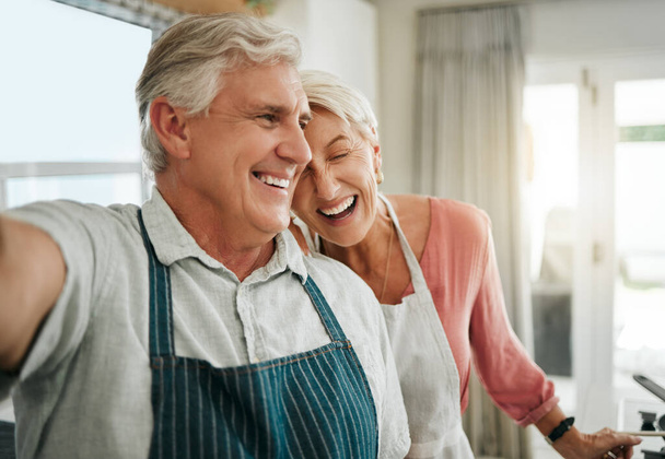 Ηλικιωμένοι, ζευγάρι και selfie στο σπίτι ευχαριστημένοι, ενώ το μαγείρεμα, το ψήσιμο ή τον καθαρισμό μαζί σε αστεία στιγμή. Μεγαλύτερος άνδρας, γυναίκα και συνταξιοδότηση γελάνε στην κουζίνα, δένουν και χαμογελούν, για να κάνουν φαγητό και αστείο. - Φωτογραφία, εικόνα