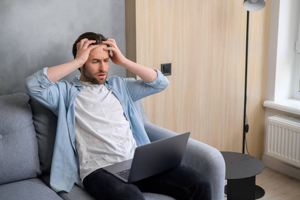 Young caucasian male worker received email with bad news, sitting at the sofa with an open laptop. Businessman lost his business, office man got faired, feeling scared, stressed and frustrated - Fotó, kép