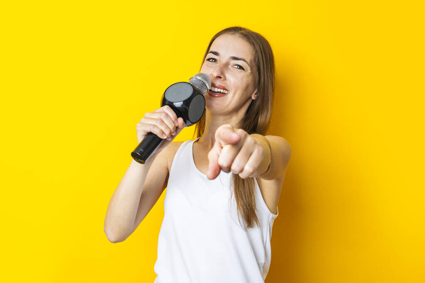 Singing young woman with a microphone points her finger forward on a yellow background. - Фото, изображение
