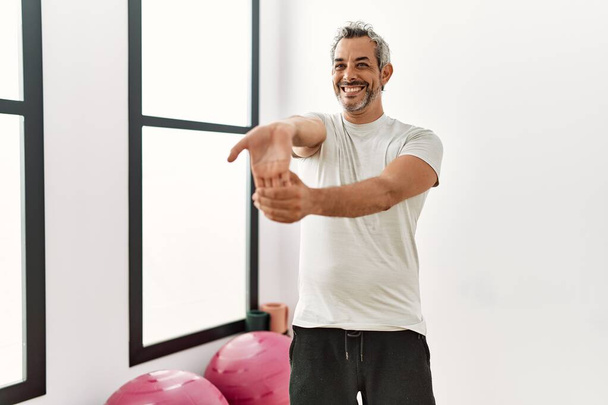 Middelbare leeftijd grijsharige man glimlachend zelfverzekerd stretching arm bij sportcentrum - Foto, afbeelding