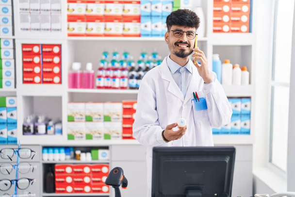 Молодий іспаномовний чоловік фармацевт розмовляє на смартфоні тримаючи таблетки в аптеці
 - Фото, зображення