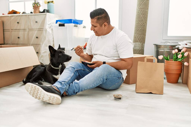 Young latin man eating taka away sushi food sitting on the floor at new home with dog. - Photo, Image