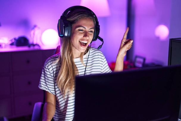 Fiatal szőke nő játszik videojátékok mosolygós boldog mutató kézzel és ujjal oldalra  - Fotó, kép