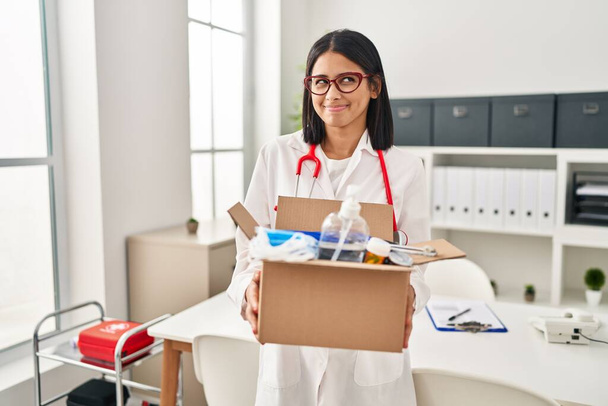 Young hispanic doctor woman holding box with medical items smiling looking to the side and staring away thinking.  - Foto, Imagen