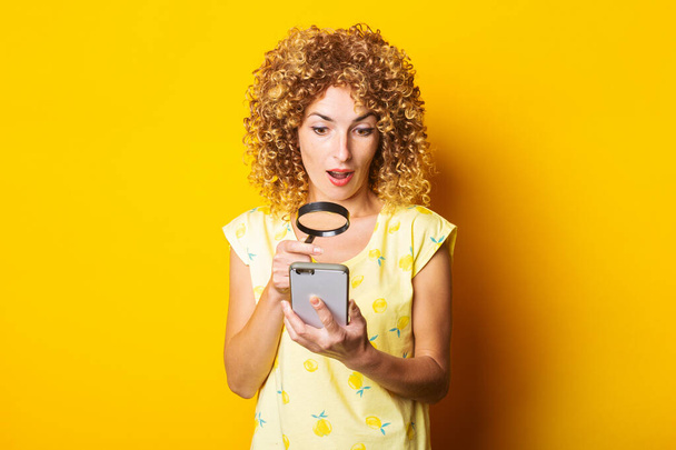 shocked curly young woman looking with a magnifying glass into the phone on a yellow background - Photo, Image