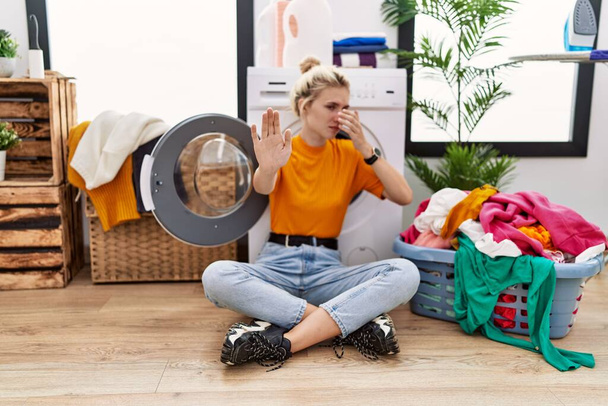 Young blonde woman doing laundry sitting by washing machine covering eyes with hands and doing stop gesture with sad and fear expression. embarrassed and negative concept.  - Photo, Image