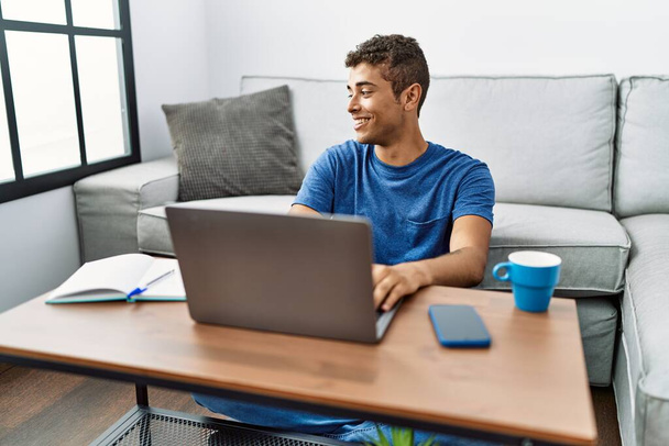 Fiatal jóképű spanyol férfi laptoppal ül a padlón, oldalra néz mosollyal az arcán, természetes kifejezéssel. nevetés magabiztos.  - Fotó, kép