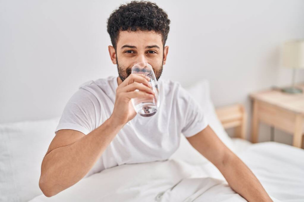 Hombre árabe joven bebiendo vaso de agua sentado en la cama en el dormitorio - Foto, imagen
