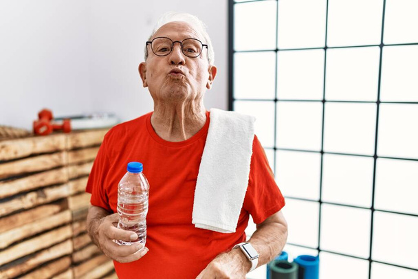 Starszy mężczyzna ubrany w strój sportowy i ręcznik na siłowni, patrzący w kamerę, dmuchający pocałunek na antenie, jest uroczy i seksowny. wyrażenie miłości.  - Zdjęcie, obraz
