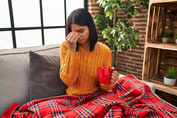 Eine junge hispanische Frau sitzt auf dem Sofa und trinkt zu Hause einen Kaffee und reibt sich müde Nase und Augen und spürt Müdigkeit und Kopfschmerzen. Konzept für Stress und Frustration.  - Foto, Bild