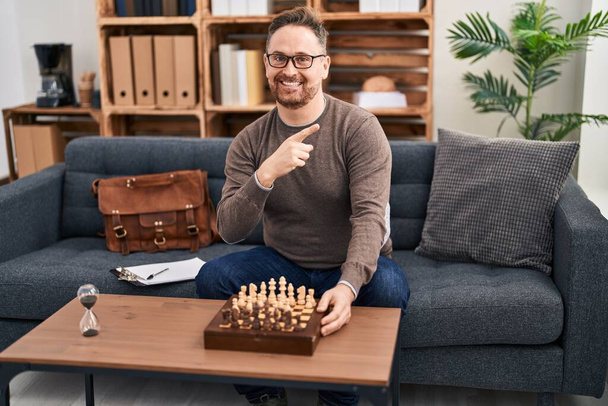 Orta yaşlı, beyaz bir adam kanepede oturmuş satranç oynuyor. Gülümsüyor, mutlu bir şekilde elini ve parmağını yana doğru gösteriyor.  - Fotoğraf, Görsel