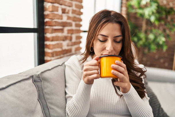 Young hispanic woman drinking coffee sitting on sofa at home - Foto, imagen