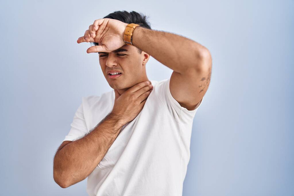Hispanischer Mann, der vor blauem Hintergrund steht und die Stirn berührt wegen Krankheit und Fieber, Grippe und Erkältung, Viruserkrankung  - Foto, Bild
