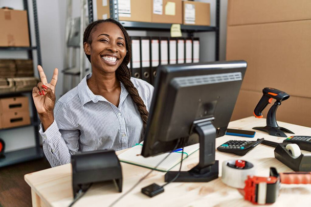 Африканская женщина, работающая в электронной коммерции малого бизнеса, улыбается, глядя в камеру, показывая пальцы, делающие знак победы. Номер два.  - Фото, изображение