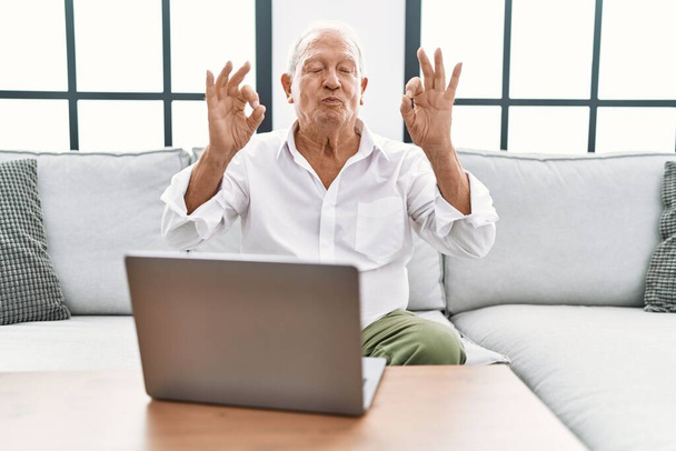 Старший мужчина, сидя дома с ноутбуком на диване, расслабляется и улыбается с закрытыми глазами, делая жест медитации пальцами. Концепция йоги.  - Фото, изображение