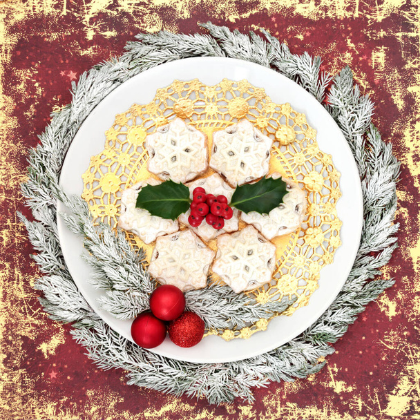 Festive Christmas traditional mince pies on white plate with winter holly, snow covered juniper fir, tree decorations on grunge red background. Food design for Xmas and New Year.  - Photo, Image