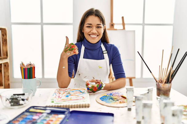 Jeune femme brune au studio d'art avec des mains peintes faisant des pouces heureux geste avec la main. approuvant l'expression regardant la caméra montrant le succès.  - Photo, image