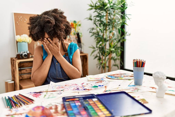 Schöne afrikanisch-amerikanische Frau mit Afro-Haarmalerei im Kunststudio mit traurigem Gesichtsausdruck, der das Gesicht mit Händen bedeckt, während sie weint. Depressionskonzept.  - Foto, Bild