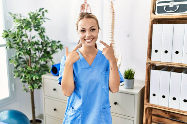 Молода біла жінка, яка працює в клініці відновлення болю, усміхається весело показуючи і вказуючи пальцями зуби і рот. концепція здоров'я зубів
.  - Фото, зображення
