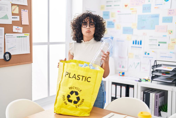 Νεαρή γυναίκα της Μέσης Ανατολής κρατώντας τσάντα ανακύκλωσης με πλαστικά μπουκάλια στο γραφείο καταθλιπτική και ανησυχούν για την αγωνία, κλάμα θυμωμένος και φοβισμένος. λυπημένη έκφραση.  - Φωτογραφία, εικόνα