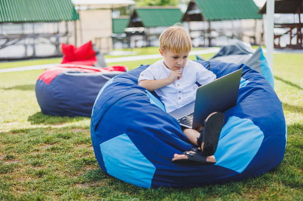 Маленький хлопчик з ноутбуком на тлі літньої трави. Хлопчик у білій футболці сидить на сумочці в парку
. - Фото, зображення