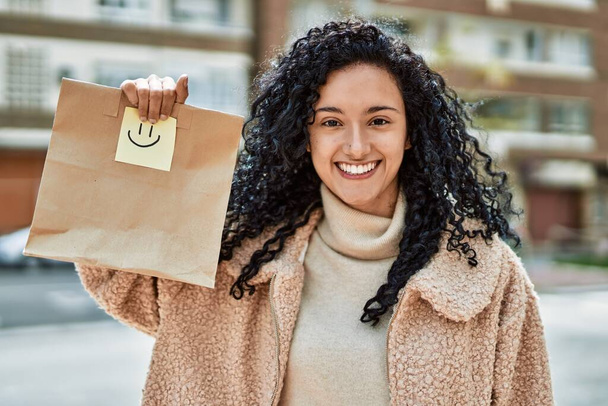Молодая латиноамериканка, уверенно улыбающаяся, держа в руках бумажный пакет на улице - Фото, изображение