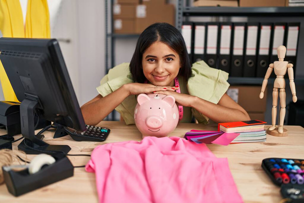 Young latin woman ecommerce business worker leaning on piggy bank at designer studio - Photo, Image