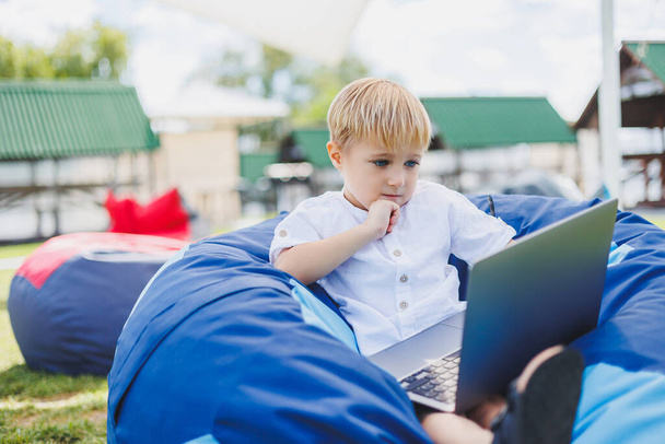Niño pequeño con un portátil en el fondo de la hierba de verano. Un chico con una camiseta blanca está sentado en una bolsa de frijoles en el parque. - Foto, imagen