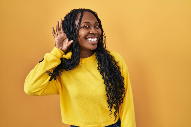 Femme africaine debout sur fond jaune souriant avec la main sur l'oreille en écoutant une rumeur ou des commérages. concept de surdité.  - Photo, image