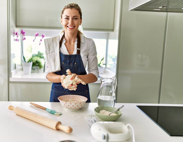 Junge blonde Frau lächelt selbstbewusst und macht Pizzateig mit Händen in der Küche - Foto, Bild