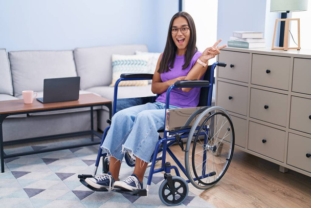 Młoda latynoska kobieta siedząca na wózku inwalidzkim w domu uśmiechnięta ze szczęśliwą twarzą mrugającą przed kamerą robiącą znak zwycięstwa. numer dwa..  - Zdjęcie, obraz