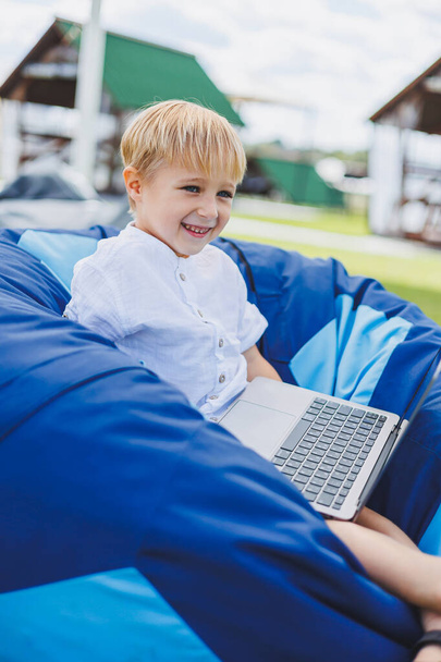 Un niño alegre está jugando en sillas coloridas en la calle. Un niño pequeño ve dibujos animados en un portátil mientras está sentado en una silla en el parque - Foto, imagen