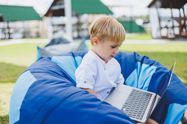 A cheerful child is playing on colorful beanbag chairs on the street. A little boy watches cartoons on a laptop while sitting on a chair in the park - Photo, Image
