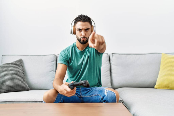 Hombre hispano guapo usando auriculares jugando a videojuegos sosteniendo el controlador apuntando con el dedo a la cámara y a ti, gesto confiado que se ve serio  - Foto, Imagen