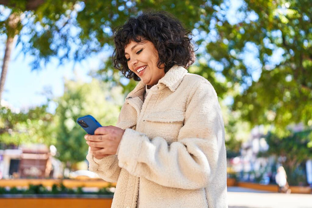 Молодая красивая латиноамериканка уверенно улыбается, используя смартфон в парке - Фото, изображение
